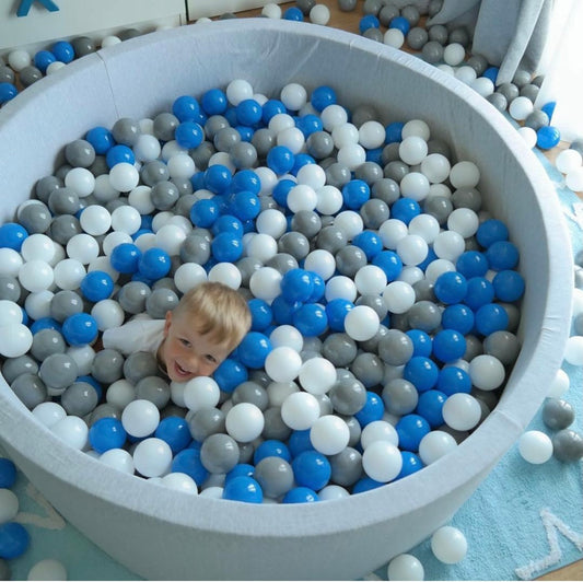 Round toddler ball pit with white, grey, and blue balls, toddler smiling at someone above the camera.