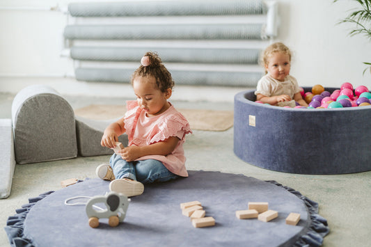 Crushed grey velvet play mat for babies, with one toddler playing and another looking off-camera, ball pit and toys in the background.