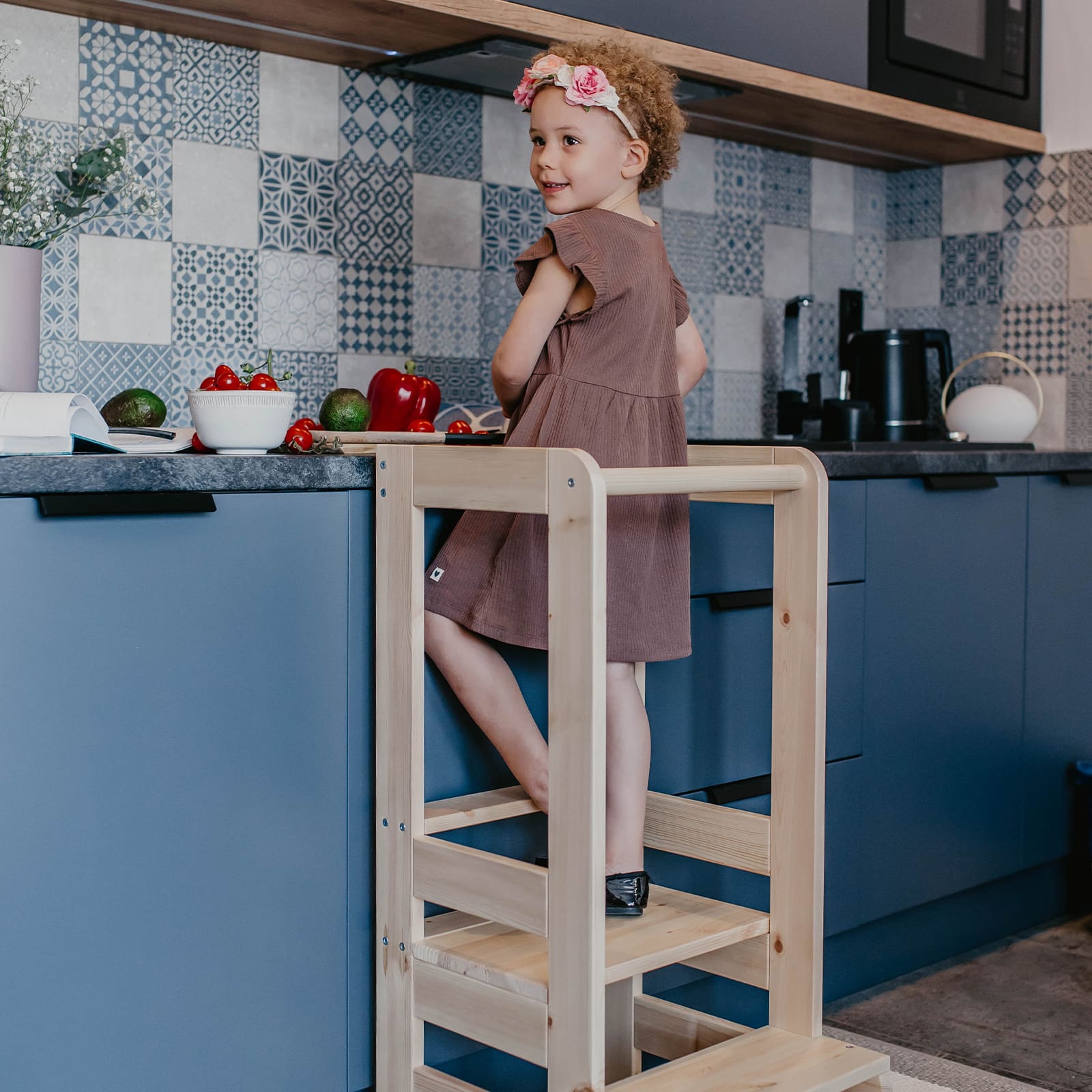 Toddler looking at the camera while helping on a kitchen helper stool in the kitchen.