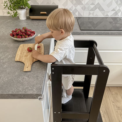 Side view of a toddler standing on a black Meowbaby kitchen helper, facing the camera.
