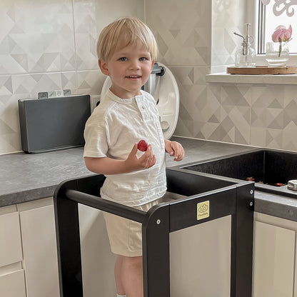 Toddler standing on a black Meowbaby kitchen helper, looking at the camera.