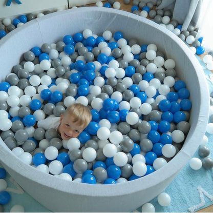 Round toddler ball pit with white, grey, and blue balls, toddler smiling at someone above the camera.