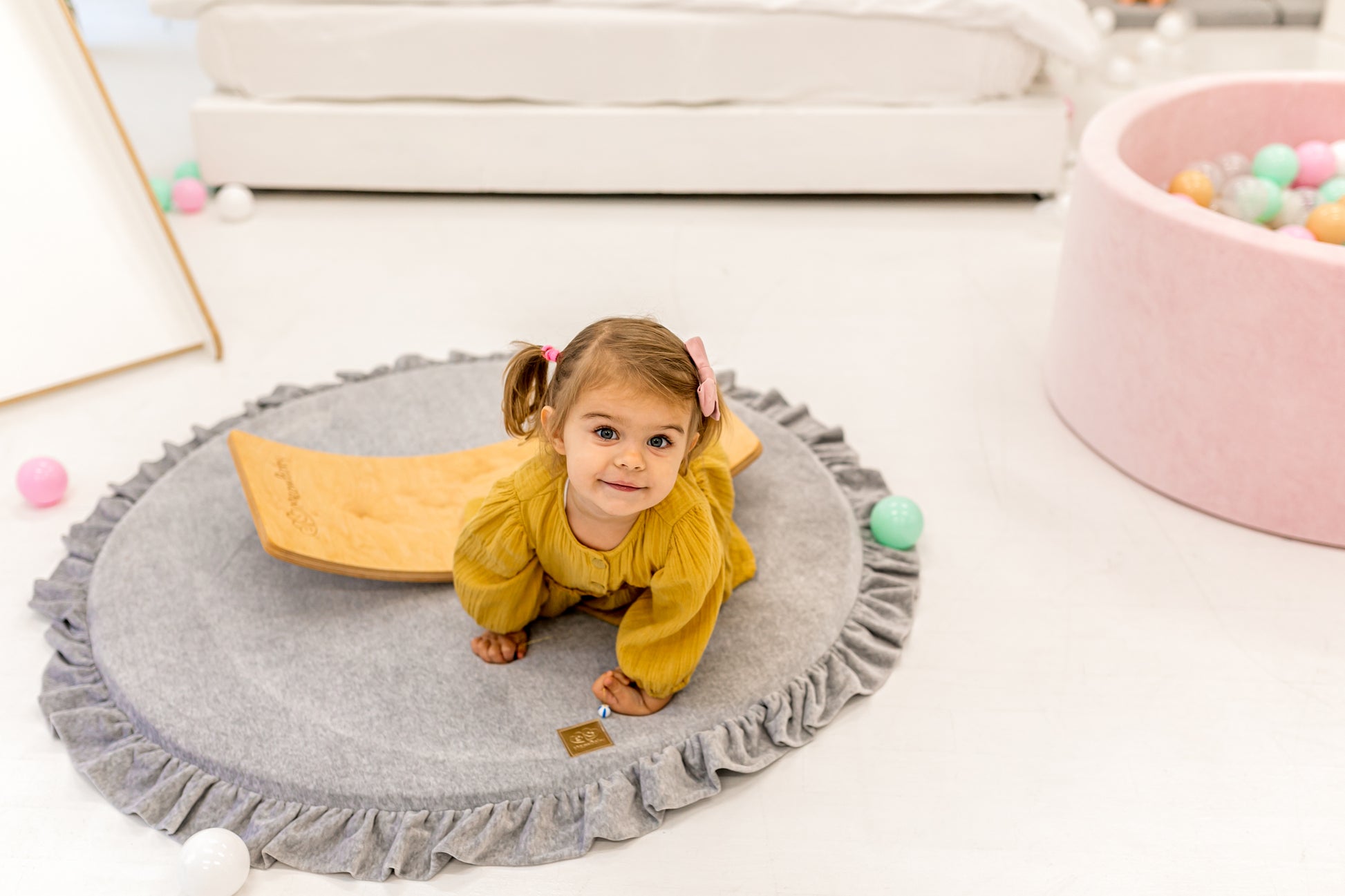 Crushed grey velvet play mat for babies, with a toddler looking at the camera and ball pit and soft play toys in the background.